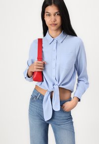 Lyseblå skjorte med knytefront, knappedetaljer, brystlomme og lange ermer, sammen med blå jeans og en liten rød veske.