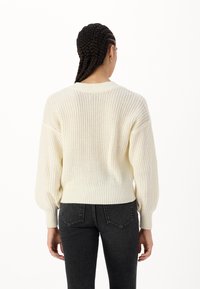 Maglione a maglia crema con una vestibilità rilassata, dotato di orli a costine e maniche a palloncino. Il tessuto testurizzato aggiunge profondità. Indossato con jeans scuri.
