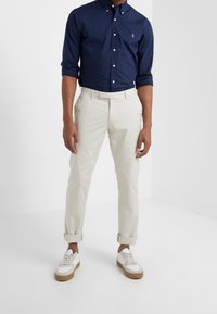 Marinflaggskjorta, krämfärgade chinos och vita sneakers med bruna sulor. Skjortan har en liten logo; chinosen är uppvikta vid ärmarna.