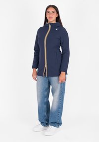 Giacca blu navy con cappuccio, zip gialla e polsini elasticizzati. Indossata sopra jeans larghi azzurri e sneaker bianche.