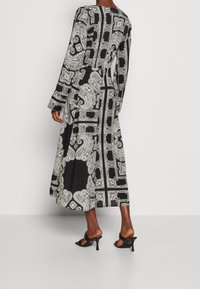Robe noire longue ornée de motifs paisley et géométriques blancs, avec manches longues et un design fluide, associée à des sandales à talons noires.