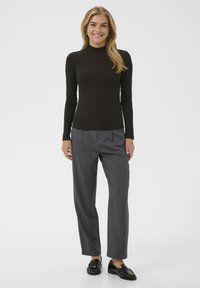 Glimlachende vrouw met lang blond haar, gekleed in een zwarte coltrui, grijze broek en zwarte instappers, staande tegen een effen achtergrond.