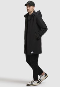 Abrigo negro impermeable con capucha, que cuenta con cierre de cremallera y bolsillos. Lleva puesto sobre una camiseta negra, combinado con pantalones cargo negros y zapatillas.