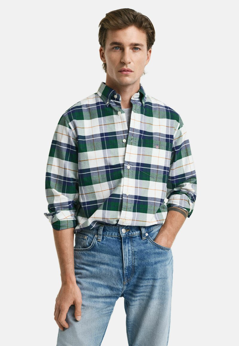 Camisa de cuadros en verde, azul marino y blanco con acentos en naranja. Cuello button-down, mangas largas enrolladas y un bolsillo en el pecho. Llevada con jeans de color azul claro.