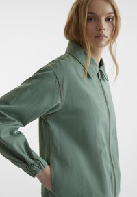 Veste zippée vert clair en tissu doux avec un col, des poignets élastiques et des détails de couture subtils. Design décontracté pour un port polyvalent.