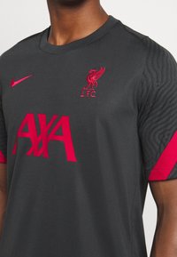 Szary sportowy trykot z czerwonymi akcentami. Posiada kołnierzyk, logo Liverpoolu, centralny nadruk "AXA" oraz wzorzyste rękawy.