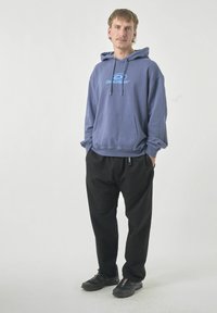 Blaue Baumwollkapuzenjacke mit einer Fronttasche und Kordelzügen, kombiniert mit lockeren schwarzen Hosen und schwarzen Sportschuhen.