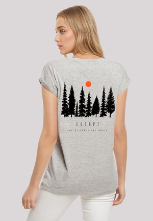 Blonde woman wearing a gray t-shirt with black pine trees and an orange sun graphic and "ESCAPE AND DISCOVER THE WORLD" text.