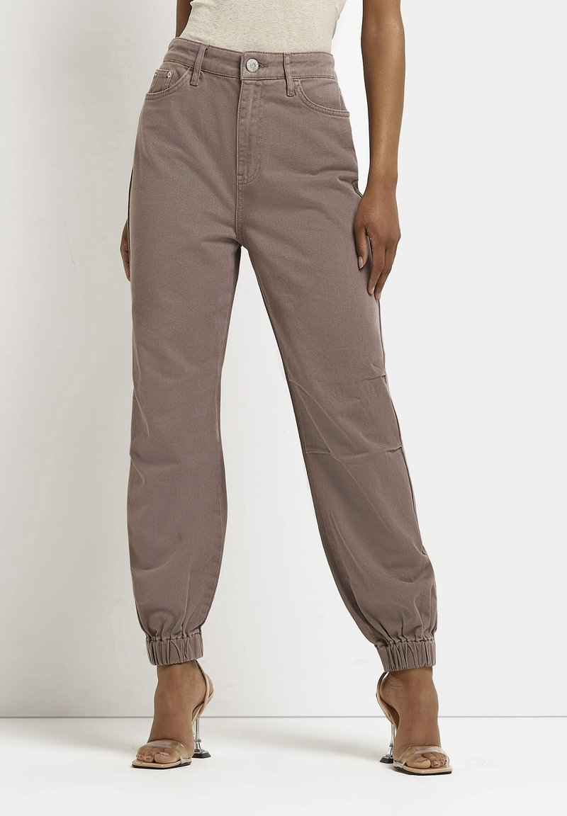 Pantalones de tiro alto color topo con puños elásticos, combinados con una camiseta de tirantes beige y sandalias de tacón alto con tiras transparentes sobre fondo blanco.