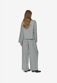 Femme avec de longs cheveux ondulés portant un costume gris ample avec un pantalon à jambes larges et des chaussures à talons noirs, se tenant de dos sur un fond blanc.