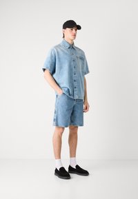 Camisa de mezclilla de manga corta en azul claro con botones y bolsillo, combinada con pantalones cortos de mezclilla a juego. Una gorra negra y unos zapatos slip-on realzan el conjunto.