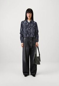 Nainen seisoo päällään sininen kuvioitu lyhyt takki, leveälahkeiset mustat farkut, mustat kengät ja pitää mustaa olkikassia sivussaan.