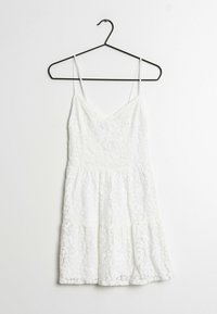 Weißes Spitzenkleid an einem schwarzen Kleiderbügel, mit schmalen Trägern, einem V-Ausschnitt, einer taillierten Oberseite und einem gestuften Saum.