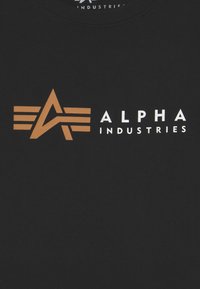 Juodas medvilninis marškinėliai su dideliu rudu ir baltu logotipu, kuriame pavaizduota abstrakti žvaigždė, o žemiau esantis užrašas "ALPHA INDUSTRIES".