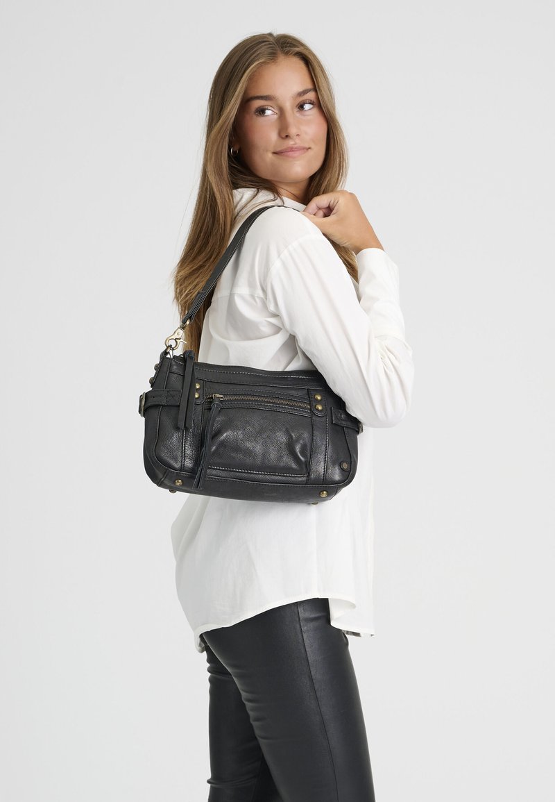 Jeune femme portant une chemise blanche et un pantalon en cuir noir porte un sac à bandoulière en cuir noir, regardant par-dessus son épaule.