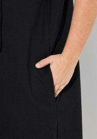 Schwarzes, strukturiertes Kleid aus Stoff mit einer Seitenleine, in die eine Hand gesteckt ist. Das Design betont Einfachheit und Funktionalität.
