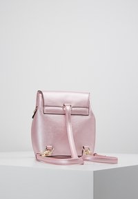 Vaaleanpunainen metallinhohtoinen reppu, jossa on sileä pinta, läppäkiinnitys, säädettävät olkahihnat ja kullanväriset metalliosat.