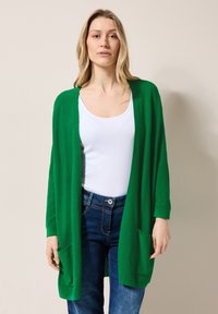 Grön öppen cardigan med långa ärmar, tillverkad av stickat material. Har två framfickor och en avslappnad passform över en vit topp och jeans.