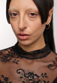 Nahaufnahme einer Person mit mittlerem Hautton, die ein durchsichtiges schwarzes Spitzentop mit Blumenmuster trägt. Goldene Creolen runden den Look ab.