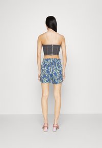 Femme vue de dos portant un haut court gris sans bretelles, un short bleu à motif cachemire, et des sandales à talons blocs roses sur fond clair.