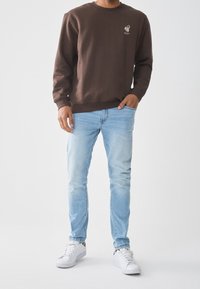 Sudadera marrón con cuello redondo y un pequeño diseño de logotipo, combinada con jeans de mezclilla azul claro y zapatillas blancas.