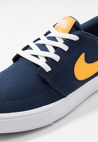 Laivastonsininen kangaskengät valkoisilla nauhoilla, keltaisella Nike-logolla, tasapohjalla ja teksturoidulla pinnalla pitävyyden ja kestävyuden takaamiseksi.