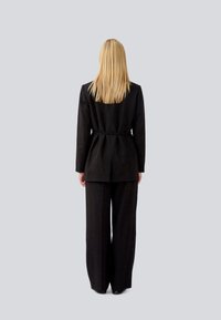 Giacca da abito nera su misura e pantaloni a gamba larga, con vita aderente, linee pulite e una texture liscia. Vista posteriore su uno sfondo grigio chiaro.