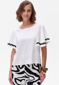 Biała bluzka z krótkim rękawem o okrągłym dekolcie, z czarnym falbanym wykończeniem na rękawach. Gładka tekstura, luźny krój. Zestawiona ze wzorzystymi spodniami.