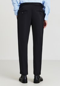 Pantaloni neri su misura con taglio dritto, dotati di due tasche posteriori e dettagli con bottoni, indossati con scarpe nere e una camicia azzurra chiara.