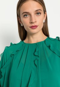 Blusa verde com um decote alto, detalhes em babados e tecido transparente. Modelo usa brincos de flores. Textura suave e cor vibrante.