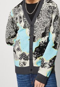 Gilet à boutonning en tissu tricoté avec un motif abstrait multicolore incluant du bleu clair, du noir et de la crème. Bordure et poignets côtelés gris.