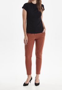 Černý top s krátkým rukávem a přiléhavým střihem, ve spojení s úzkými kalhotami v cihlové barvě, které mají šikmé švy a hladkou texturu. Černé vysoké podpatky.