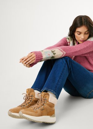 Bottes à chevilles en daim marron avec doublure en fourrure texturée, lacets épais et semelle blanche épaisse, accompagnées d'un jean en denim bleu et d'un pull à motifs roses.
