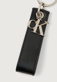 Musta nahkainen avainranneke, jossa on hopeinen "CK"-logo ja se on kiinnitetty hopeiseen avaimenperään valkoista taustaa vasten.