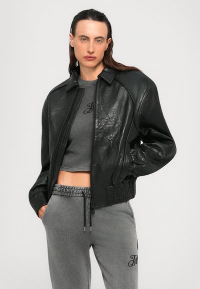 Una persona con lunghi capelli scuri indossa una giacca di pelle nera su un crop top grigio e pantaloni felpati grigi, in piedi con le mani nelle tasche.