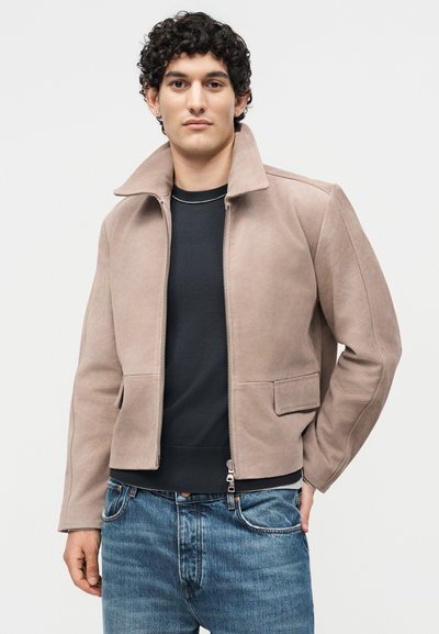 Νέος άνδρας με σγουρά μαλλιά που φοράει ένα ανοιχτό καφέ σουέντ μπουφάν πάνω από ένα μαύρο πουκάμισο και μπλε τζιν, ποζάροντας μπροστά σε απλό φόντο.