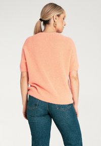 Krótki rękaw, brzoskwiniowy sweter o miękkiej fakturze. Posiada okrągły dekolt i luźny krój. W połączeniu z ciemnoniebieskimi dżinsami.