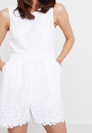 Nainen, joka pukeutunut valkoiseen, pitsireunaisella teksturoidulla olkaimettomaan haalaripukuun, kädet taskuissa, yksinkertaista valkoista taustaa vasten.