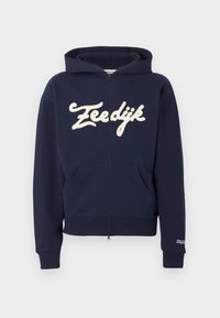 Laivastonsininen vetoketjullinen huppari, jossa etutaskut, resorit hihansuissa ja helmassa, kermaan vivahtava "Zeedijk"-teksti rinnassa ja pieni valkoinen teksti vasemman hihansuun resorissa.