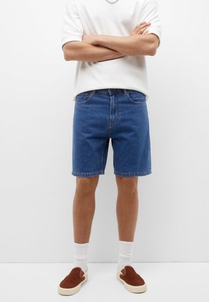 Persona che indossa pantaloncini di jeans blu, maglietta bianca, calzini bianchi e sneakers marroni senza lacci, in piedi con le braccia incrociate.