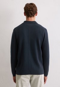 Mann, der sich abwendet und einen dunkelblauen Langarmpullover sowie hellbeige Hosen trägt, vor einem schlichten weißen Wandhintergrund.