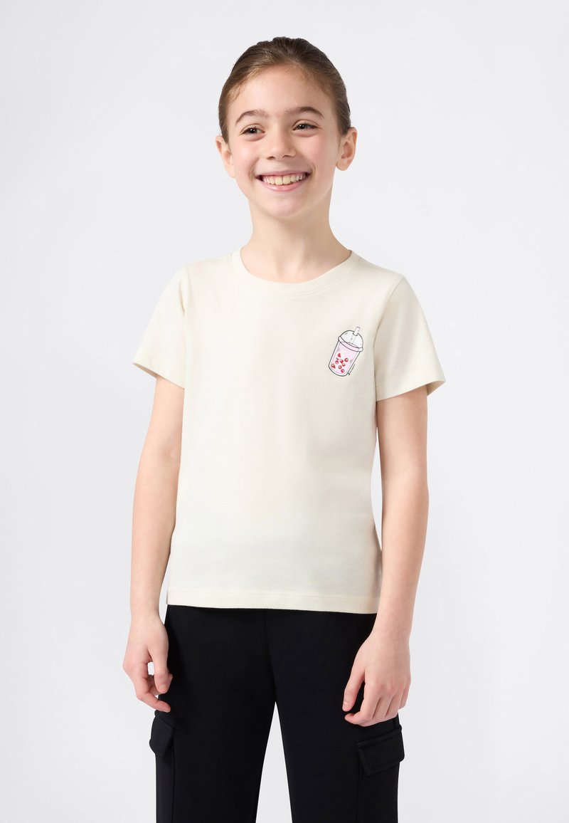 Šypsena mergaitė vilki trumpomis rankovėmis kreminę marškinėlį su burbulinės arbatos grafika ir juodomis kargo kelnėmis, stovi priešais šviesų vientisą fono.