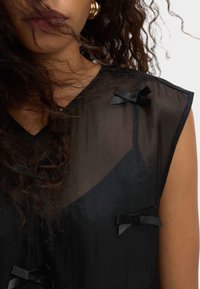 Top nero trasparente con scollatura a V, decorazioni in nastro nero e design in tessuto texturizzato. Senza maniche e con una vestibilità ampia.