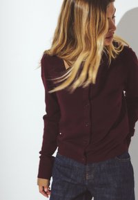 Burgundský kardigán vyrobený z jemnej látky, s predným zapínaním na gombíky a rebrovaným lemom. Spárovaný s tmavými džínsami.