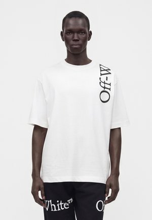 Homme à la peau foncée portant un t-shirt blanc oversize avec le texte "Off-White" sur l'épaule et un pantalon noir avec le texte blanc "Off-White".