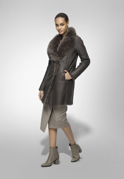 Cappotto di pelle marrone con un colletto di pelliccia voluminoso. Presenta tasche con zip, abbinato a un vestito grigio chiaro e stivaletti beige.