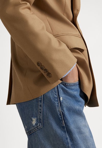 Velbloudí sako s detailem manžety na čtyři knoflíky v kombinaci s modrými džínami s opotřebovaným efektem. Sako má strukturovaný střih a hladkou texturu.
