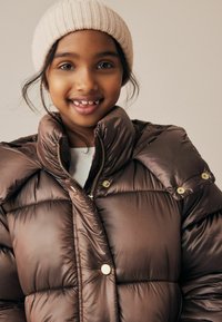 Braune Puffjacke mit hohem Kragen, goldfarbenen Beschlägen und einem gerippten, beigen Mütze. Glatte, glänzende Textur mit sichtbaren Nähten.