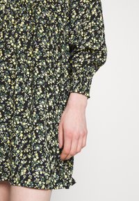 Blommig klänning med svart bakgrund, gula blommor och gröna blad, långa ärmar och rynkade manschetter, tillverkad av lättviktsmaterial.