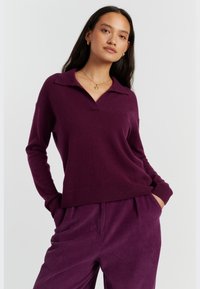 Lila gestrickter Polo-Pullover mit V-Ausschnitt, gerippten Bündchen und Saum. Kombiniert mit passenden Cordhosen, die vertikale Streifen zeigen.